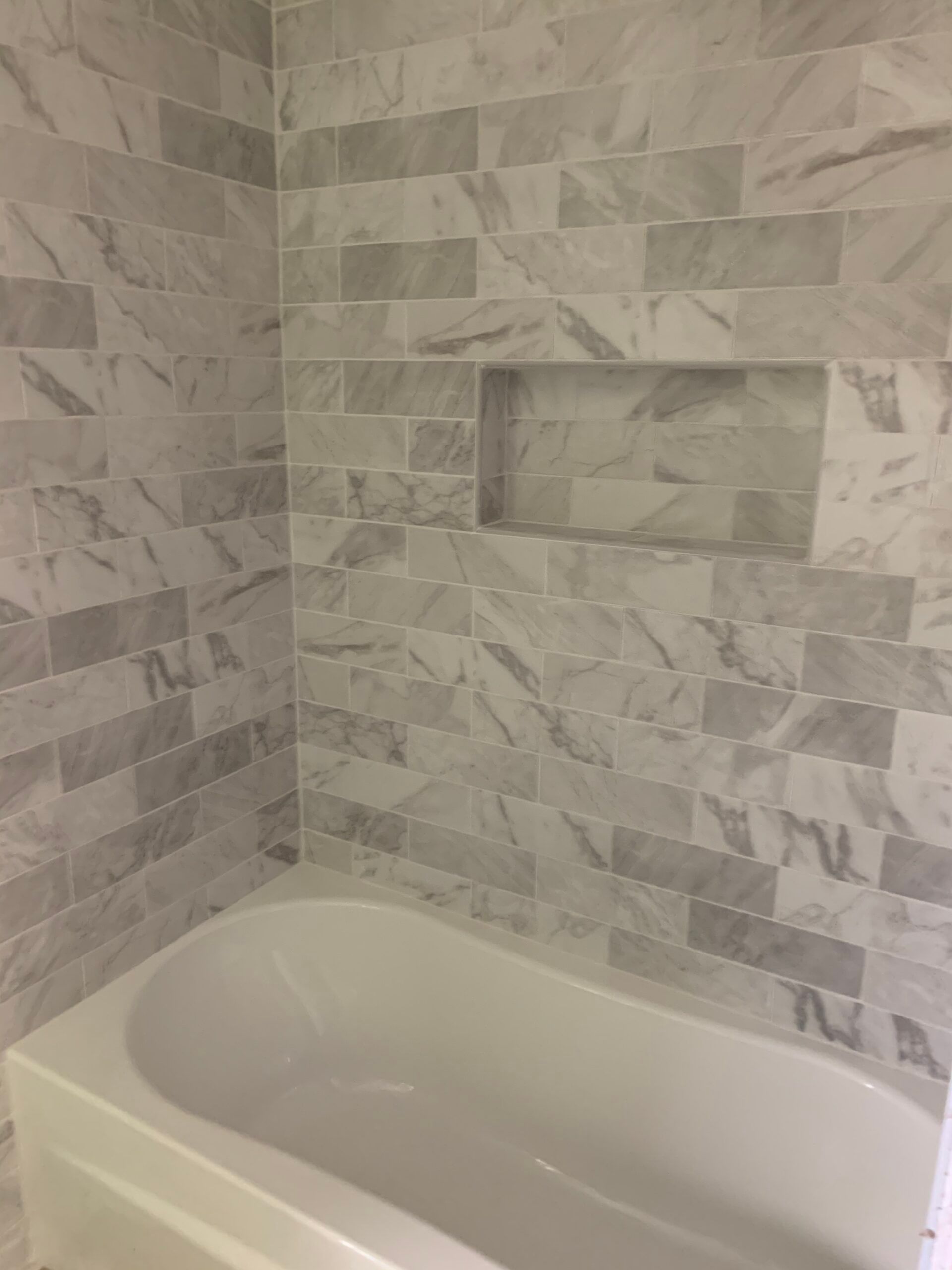White bathtub with gray tiled walls and recessed shelf.