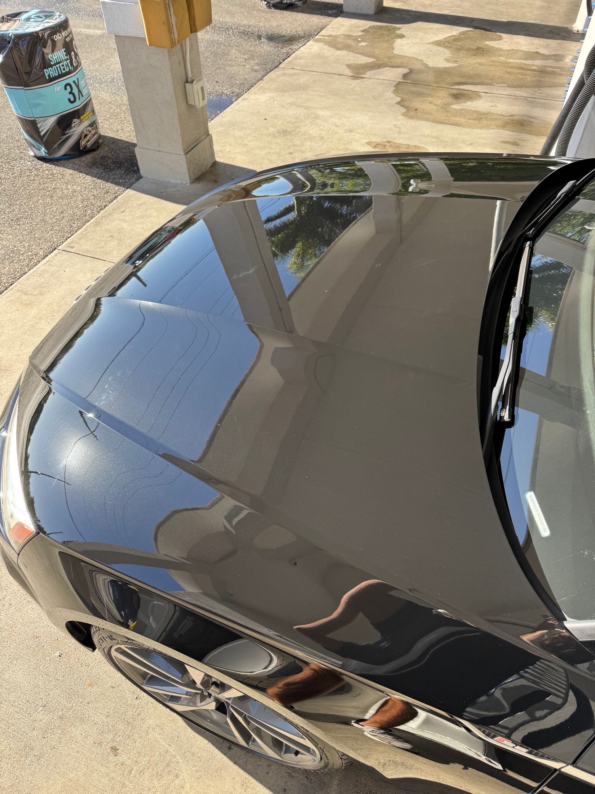 Shiny black car hood parked under a covered outdoor area, reflecting nearby buildings and sky