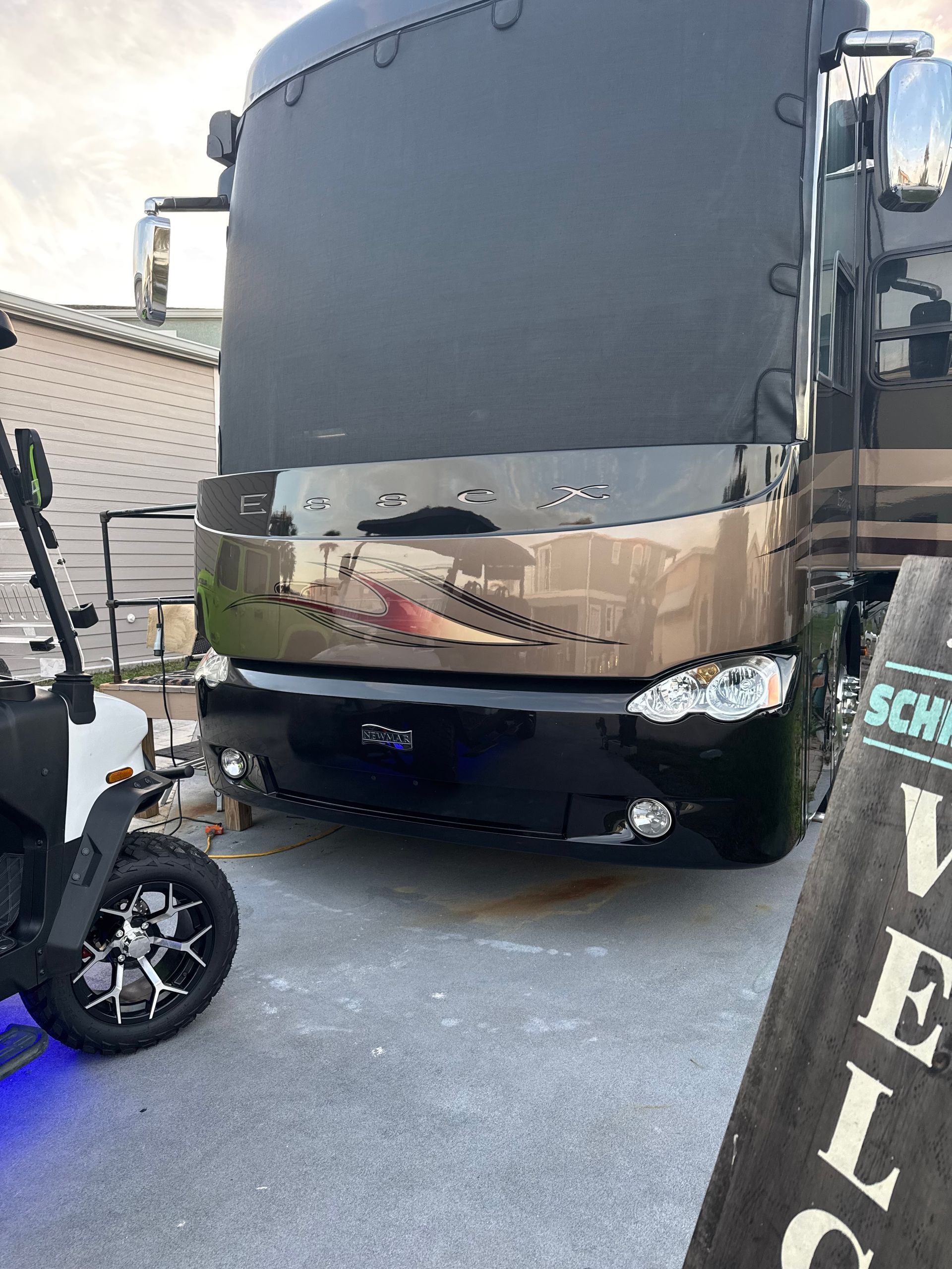 Large black bus with a reflective front parked in a narrow driveway beside a scooter and a house.
