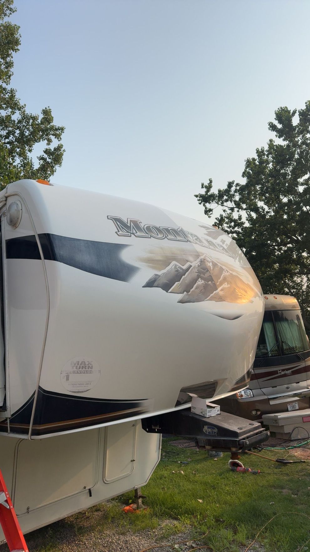 Damaged white RV with a torn front panel, parked outdoors near trees at dusk