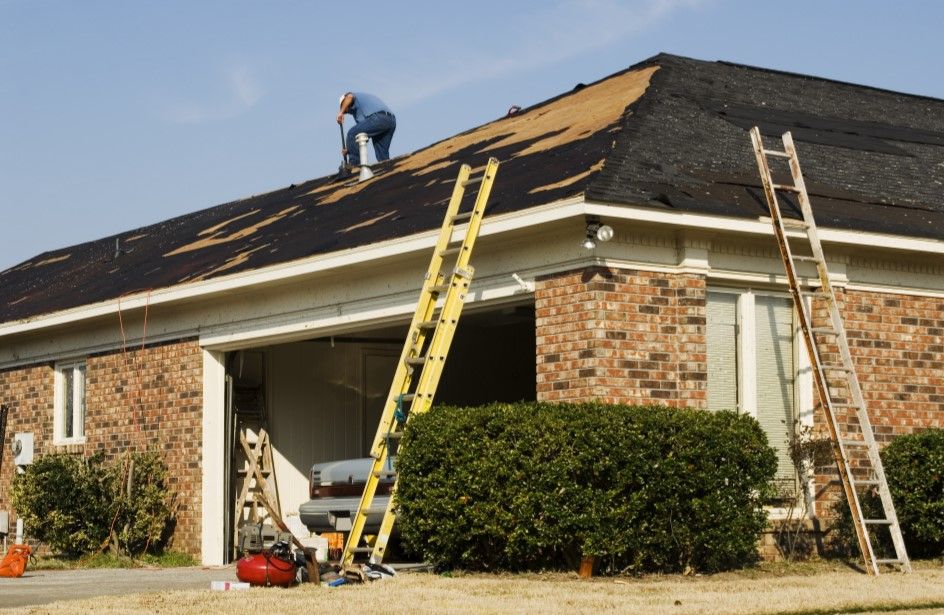 Man on a roof repairing shingles with two ladders leaning against the building under a blue sky.