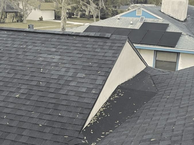 Shingle roofs of two houses, one with solar panels. Green trees and blue sky in the background.