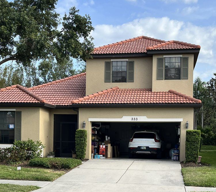 Tan two-story house with red tile roof, attached garage with a car inside, and green shutters.