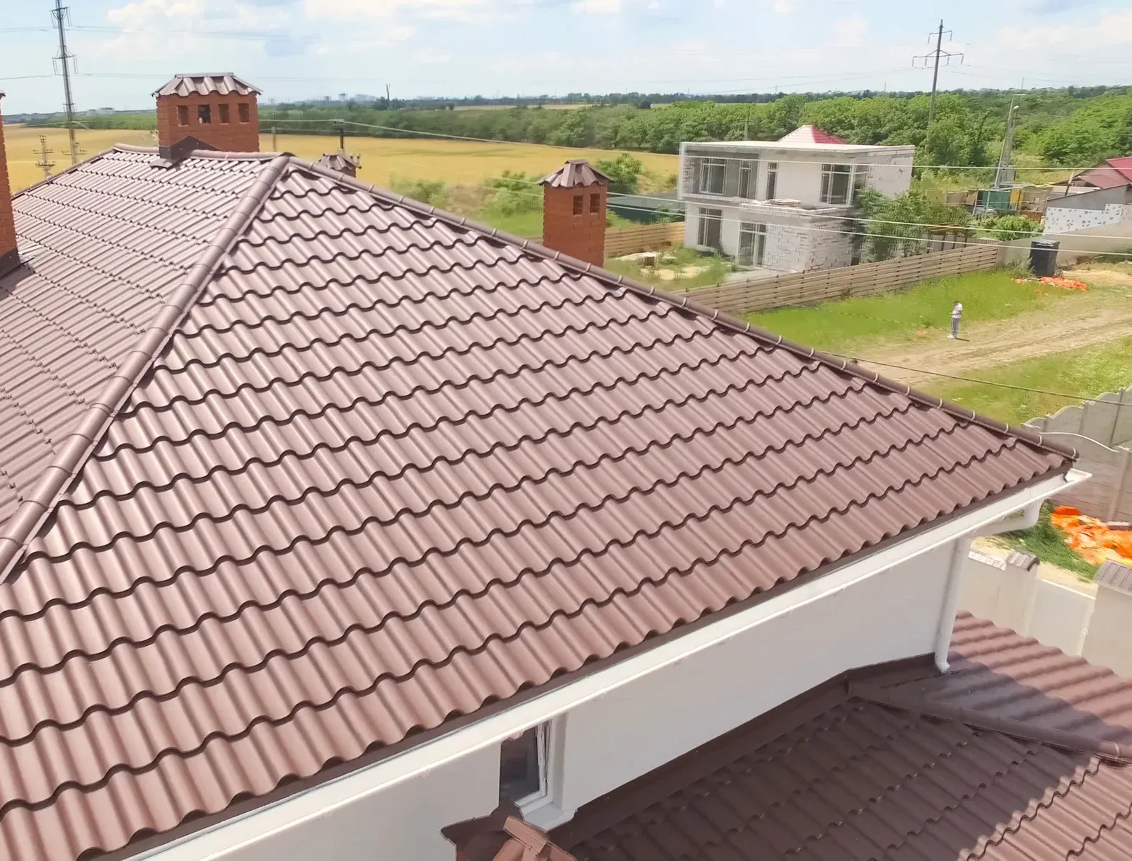 Brown metal tile roof on a house with a bright field in the background.