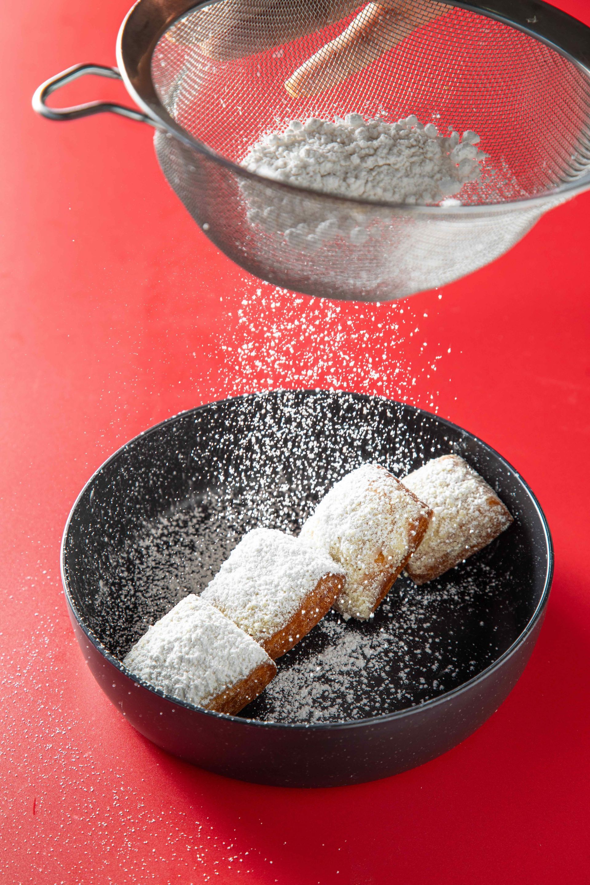 Powdered sugar sifting onto pastries in a black bowl on a red surface
