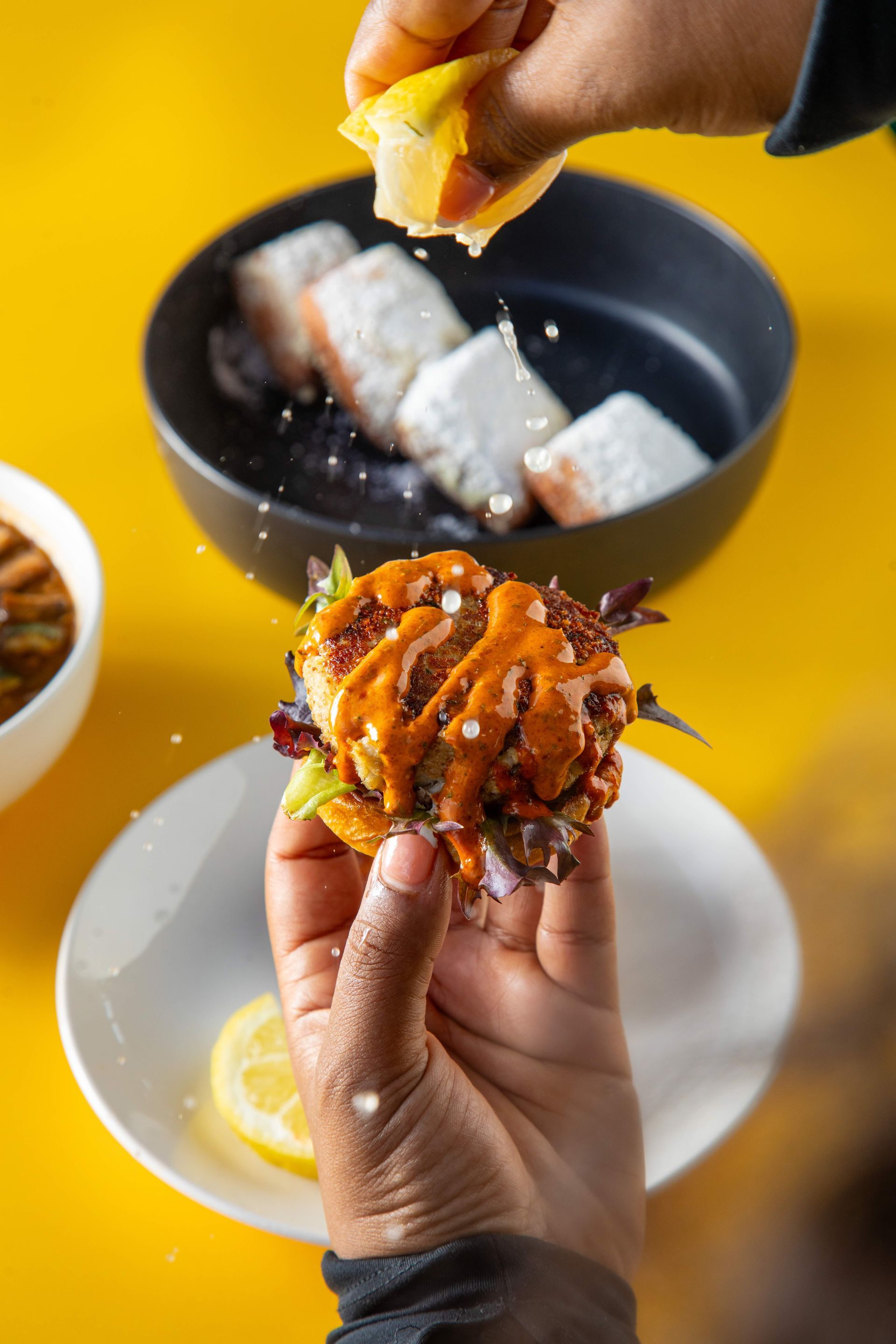 Hands holding a breaded octopus snack and lemon wedge over a bowl on a bright yellow table