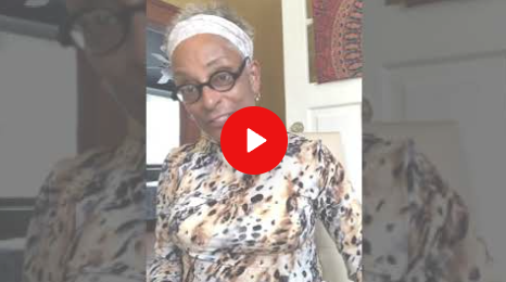Person in glasses and headwrap standing indoors, with a red play button overlay.