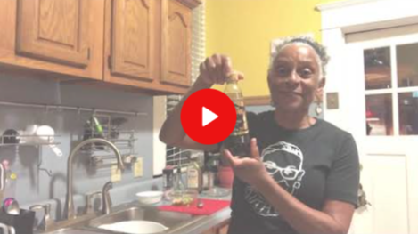 Woman in a kitchen holding up a small item near the sink, with a red play button overlay.