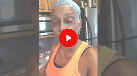 Person in an orange tank top and blue headband standing in a kitchen, with a play button overlay.