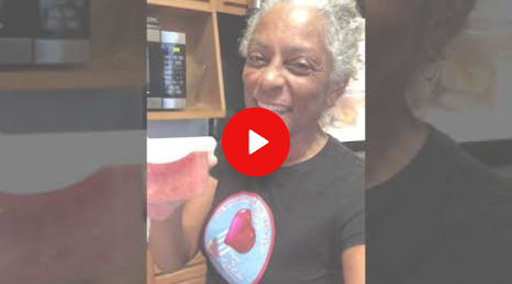Smiling person holding a red cup in a kitchen, wearing a black shirt with a heart graphic.