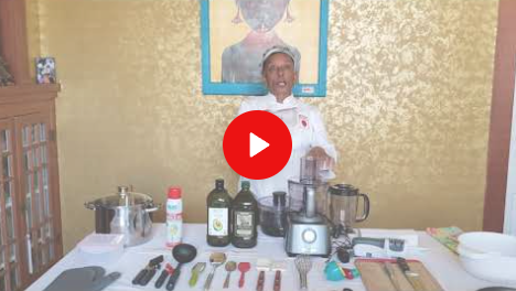 Person standing behind a table with bottles, jars, utensils, and a blender in a kitchen-like setting