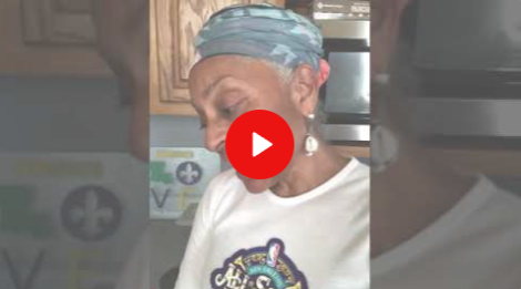 Woman in a blue headscarf and white T-shirt in a kitchen, with a red play button overlay.