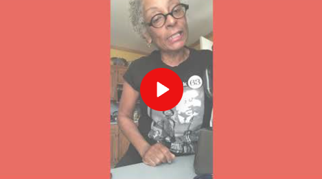 Person wearing glasses and a black graphic T-shirt, leaning toward the camera with a red play button overlay.