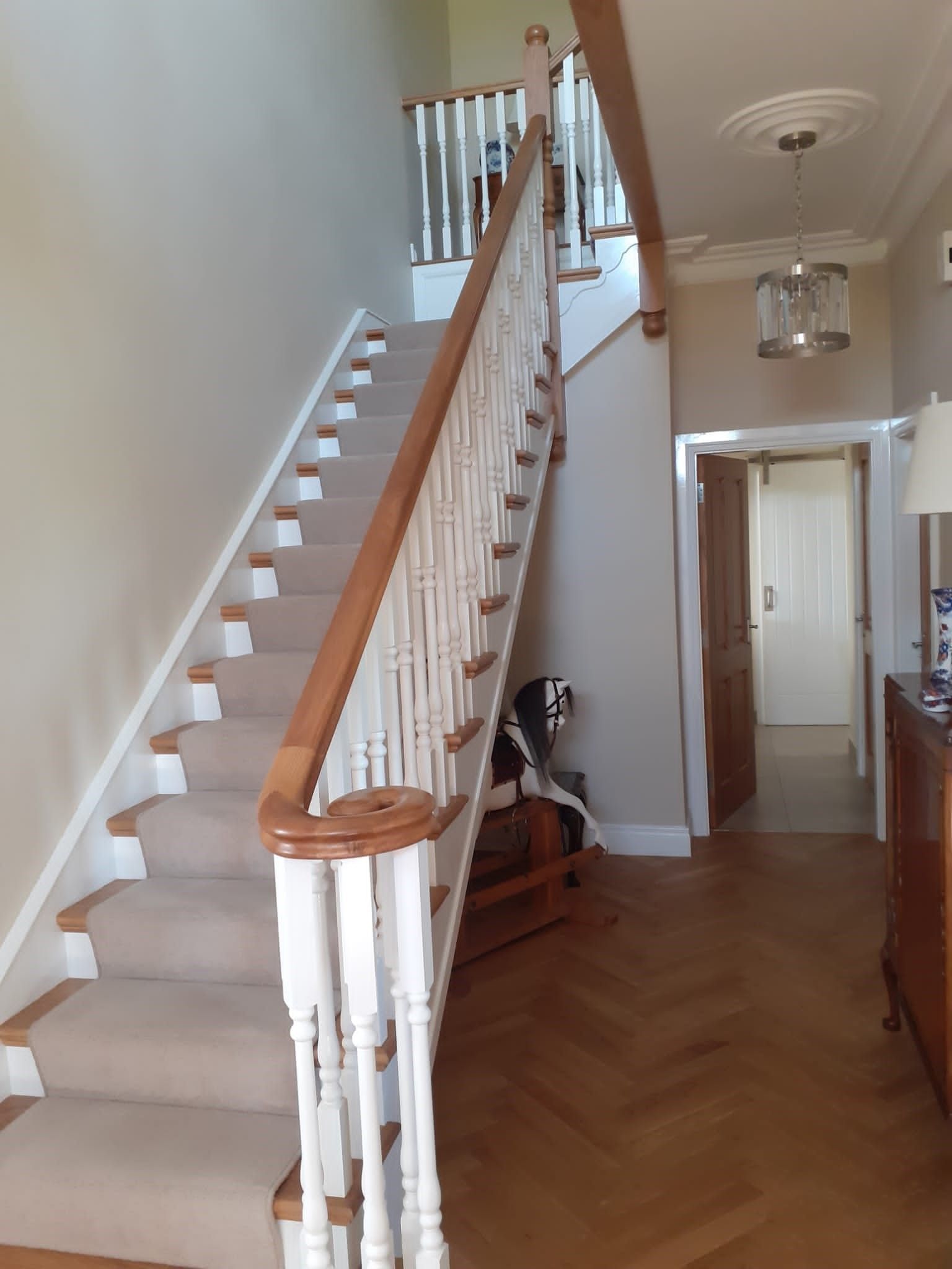 Staircase with carpeted steps and wooden banister. Herringbone wood floor leads to hallway with light fixtures.