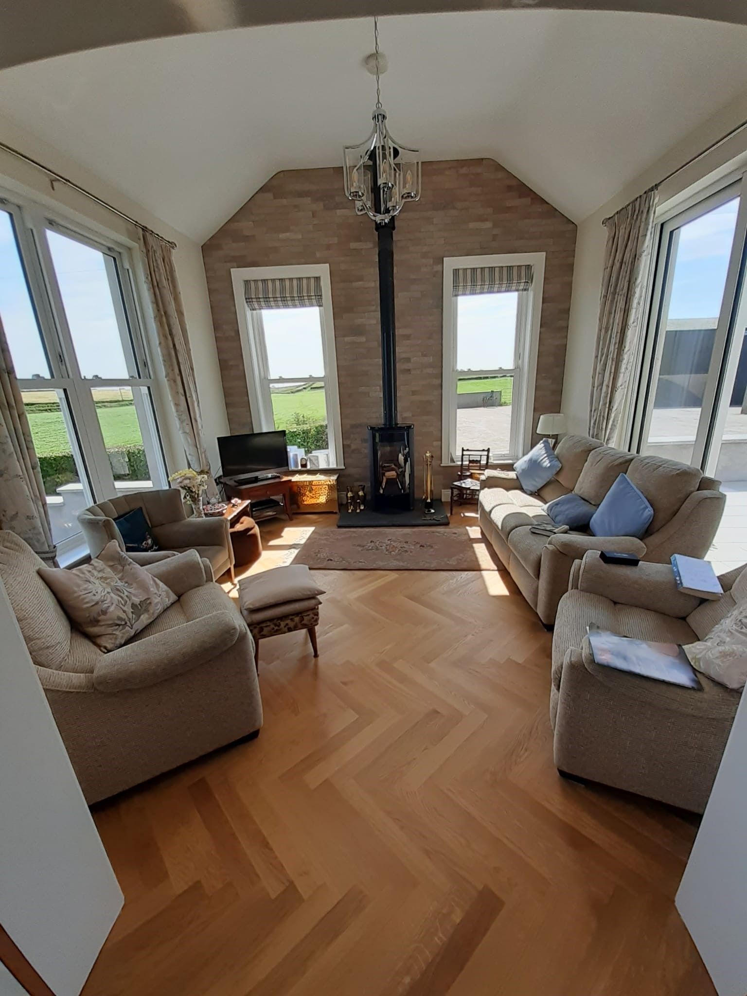 Living room with light-colored herringbone floors, sofas, fireplace, and large windows overlooking a green field.