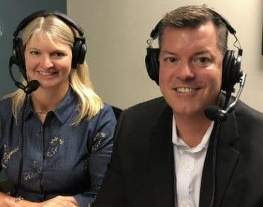 Two people wearing headsets, smiling. Woman on the left with blonde hair, man on the right in a suit.