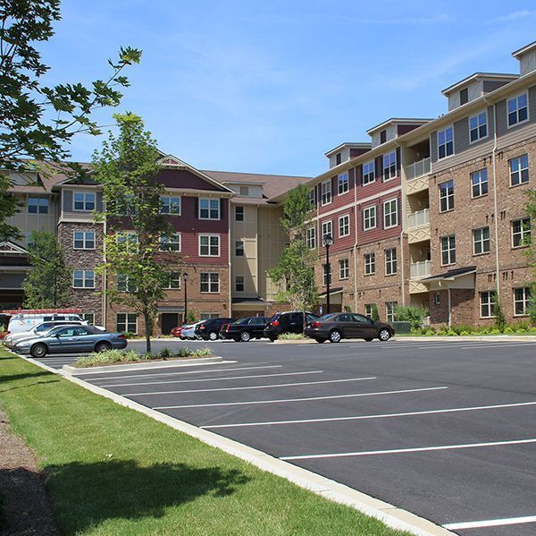 Apartment complex parking lot with parked cars, trees, and tan-and-red buildings on a sunny day