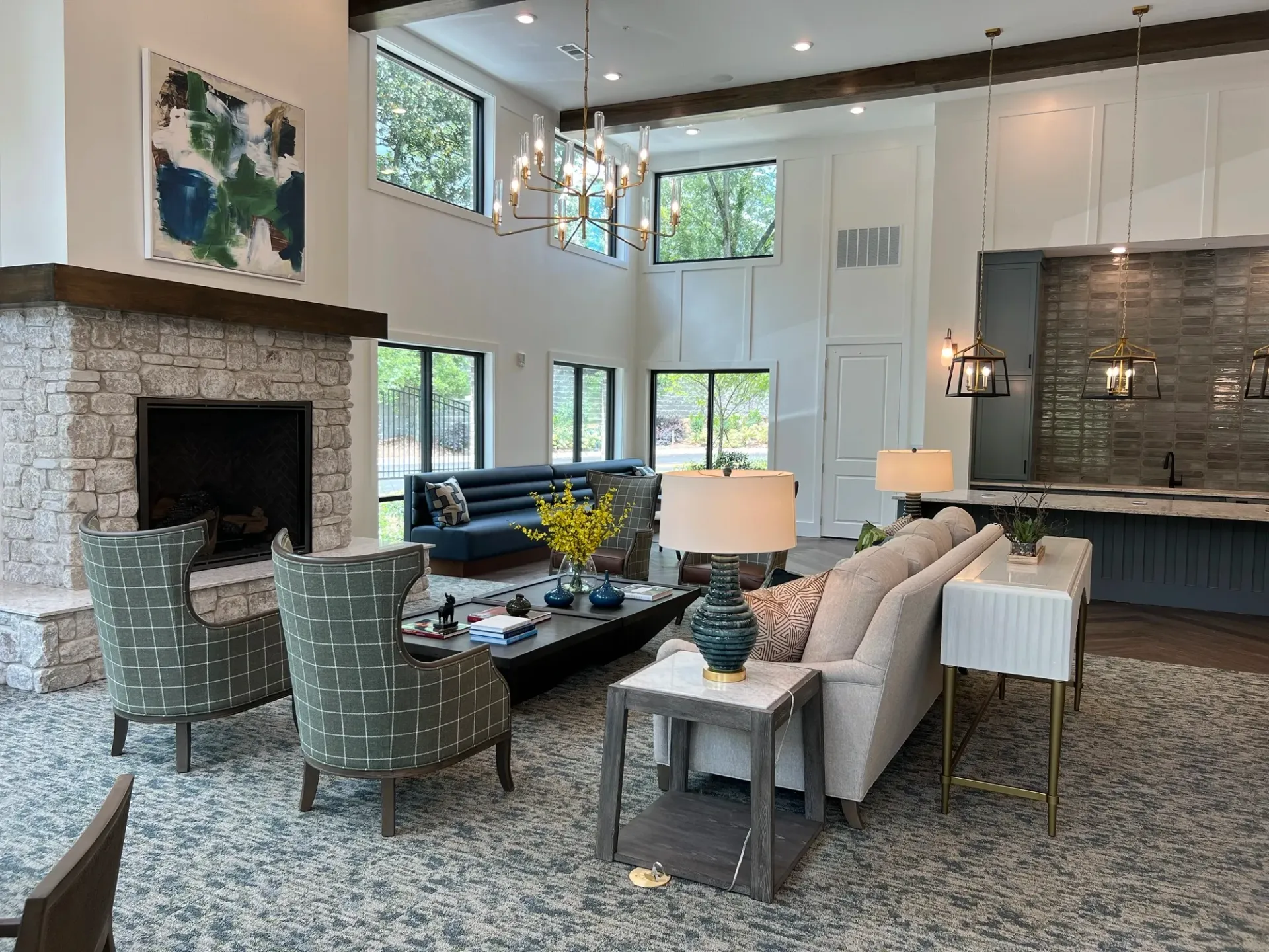Modern living room with stone fireplace, large windows, gray chairs, sofa, and kitchen island in the background