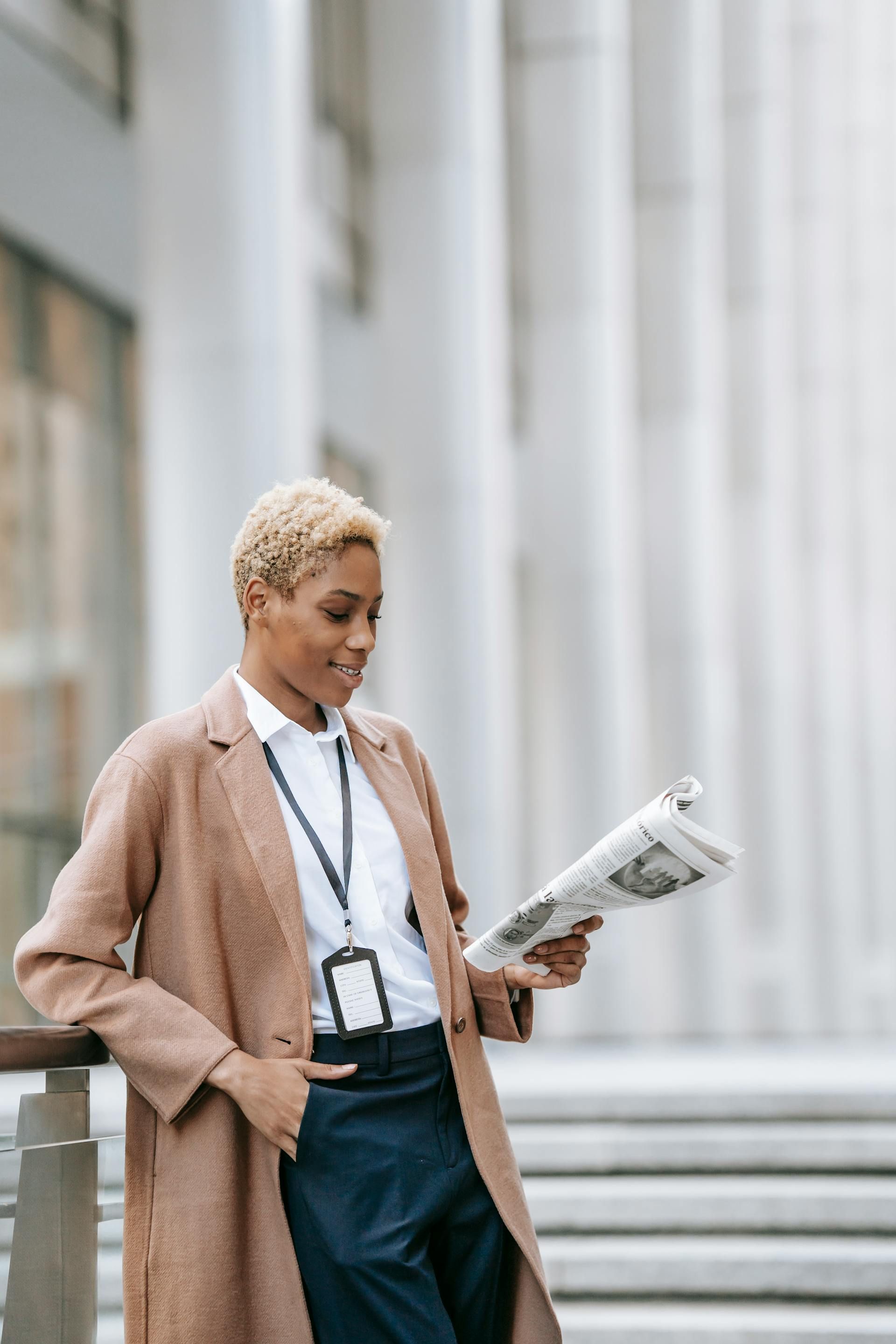 Woman in tan coat reading newspaper outdoors, leaning on railing near building.