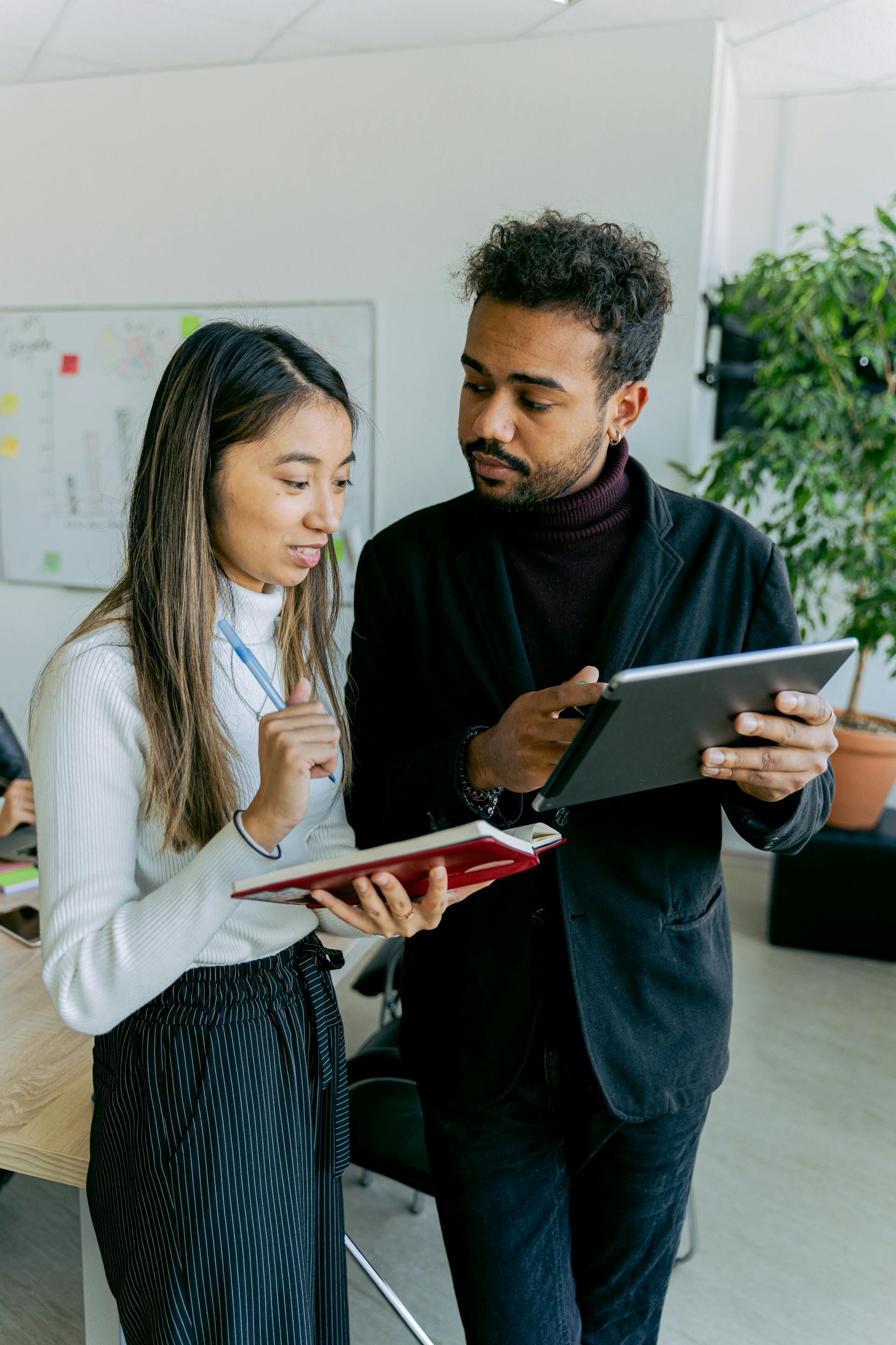 Two people in an office looking at notebooks and a tablet. Whiteboard and plant in background.