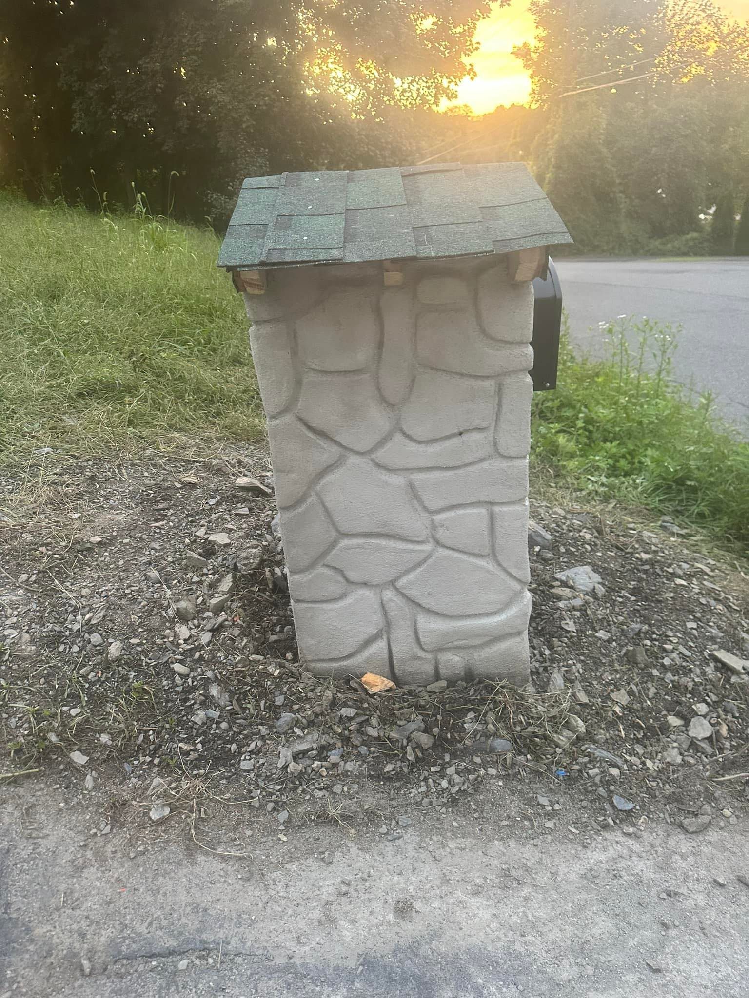 A stone mailbox with a green roof is sitting on the side of the road.