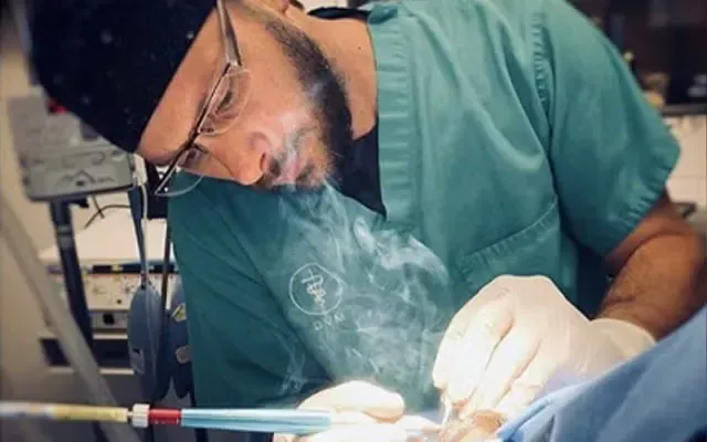 Surgeon in green scrubs and mask performing a procedure under a bright surgical light