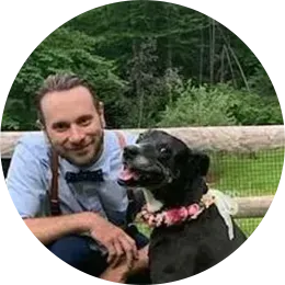 Man kneels beside a black dog wearing a pink flower collar in a grassy park, both facing the camera