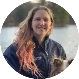 Person smiling outdoors by the water, wearing a dark jacket with long hair blowing in the wind.