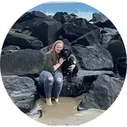 Woman sitting on rocks, holding a black dog at a rocky beach.