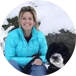 Woman in a turquoise jacket sitting in snow with a black-and-white dog, smiling at the camera.