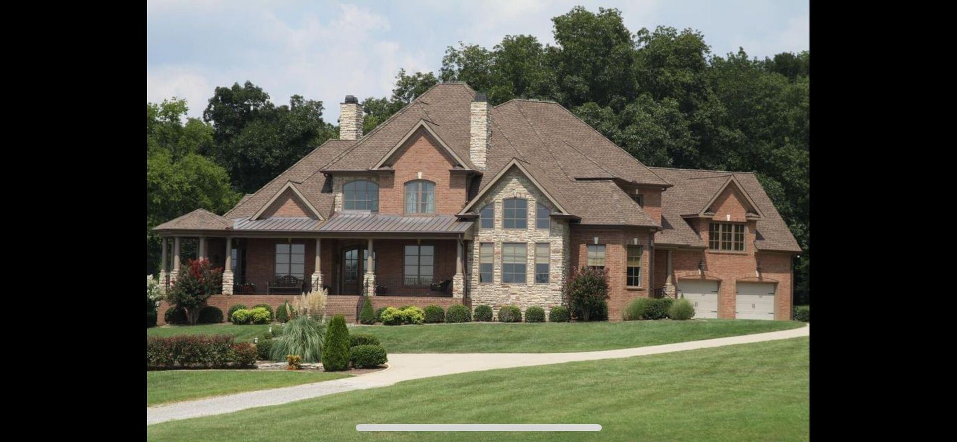 A large brick house with a covered front porch and stone accents, set against a backdrop of trees and a green lawn.