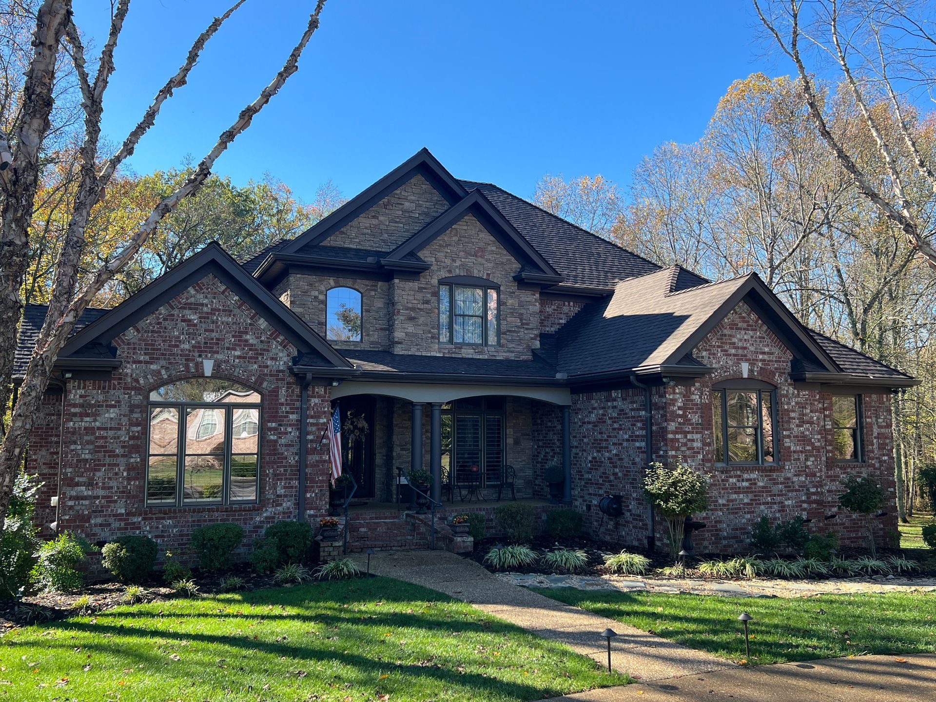 A two-story brick house with a dark gabled roof, arched front windows, and a landscaped yard under a clear blue sky.