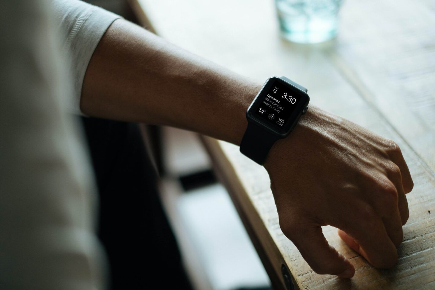 Person wearing a black smartwatch on a wooden table; the screen displays a music app.