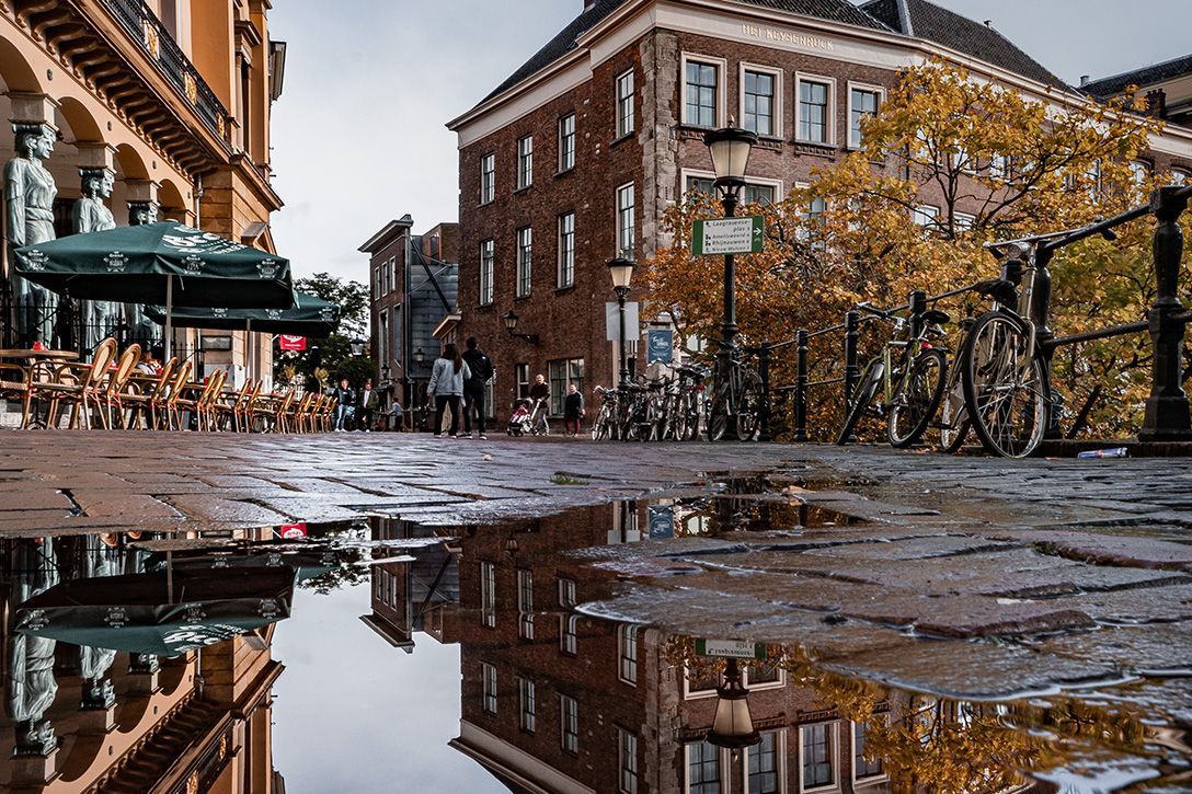 Brick street with puddles reflecting buildings and trees. Bicycles parked; people walking.