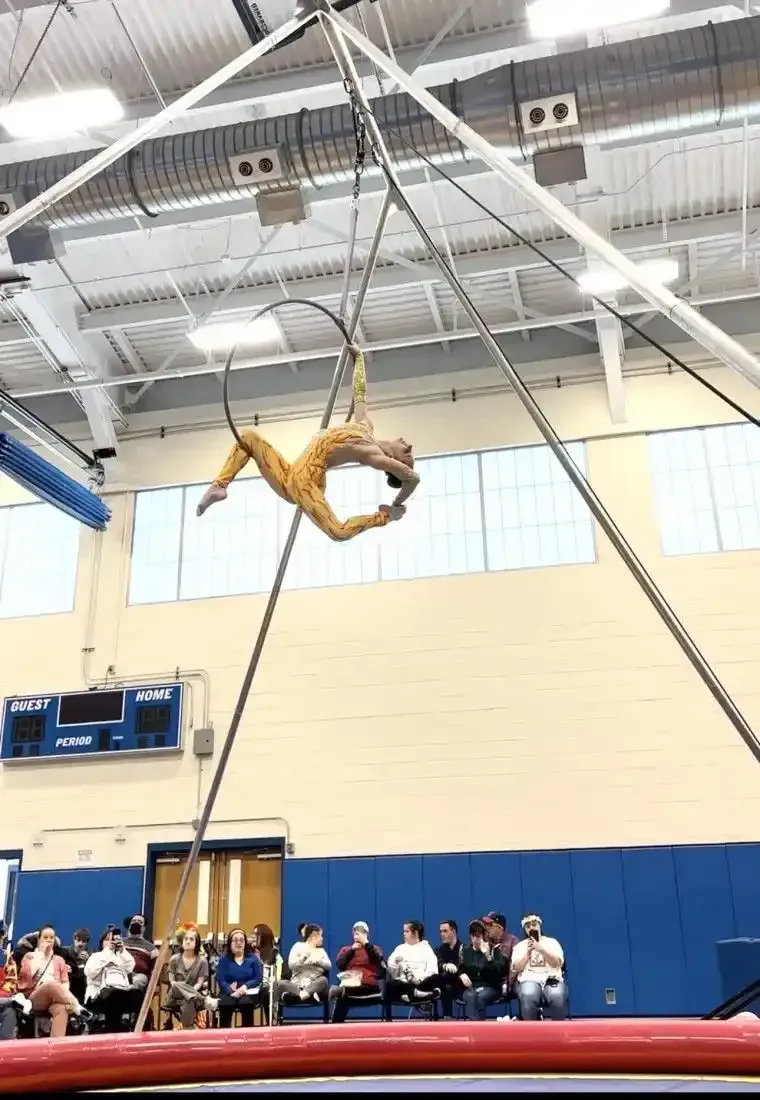 A person performs aerial acrobatics on a hoop suspended from a tall metal structure indoors.