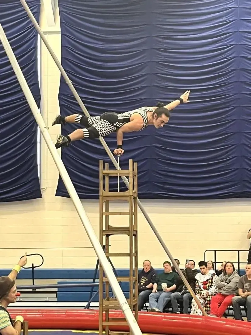 Acrobat balancing on ladder in a gym, extended horizontally two poles with audience watching behind.