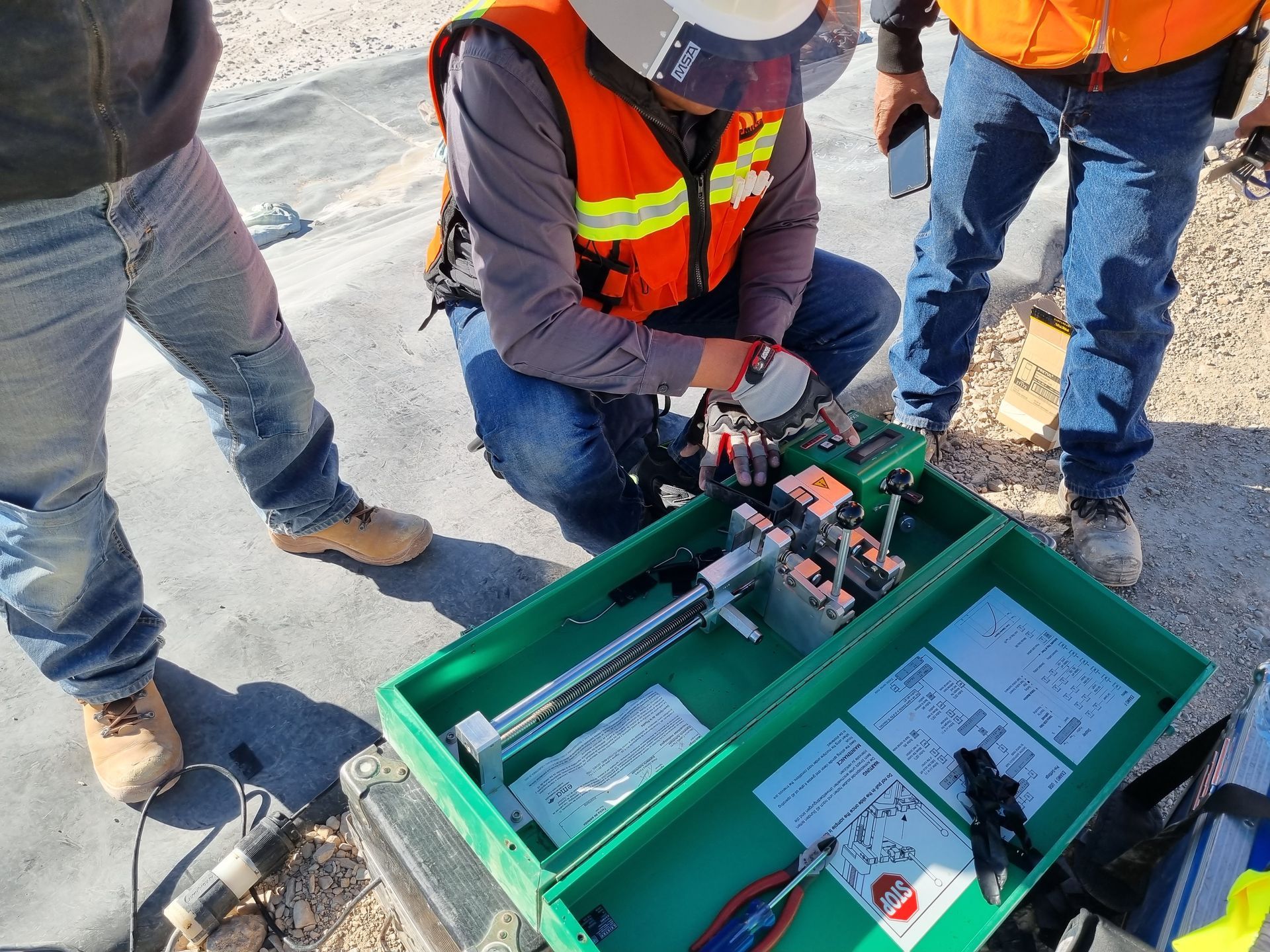 Three construction workers examining equipment in an open green box. One kneels wearing an orange vest.