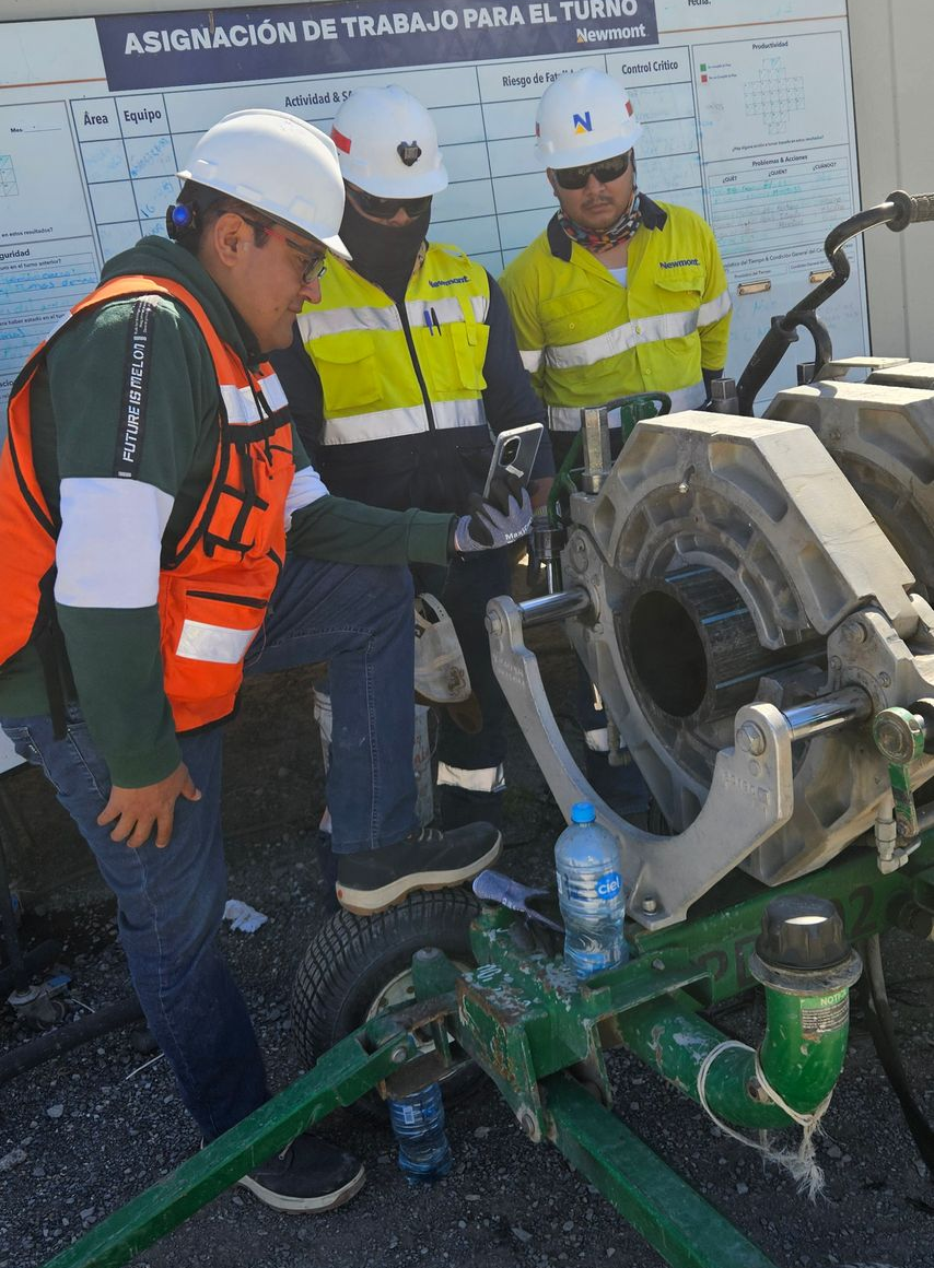 Three construction workers examining equipment outdoors, wearing helmets and vests.