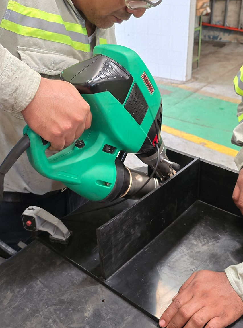 Person using a green power tool on a black container in an industrial setting.