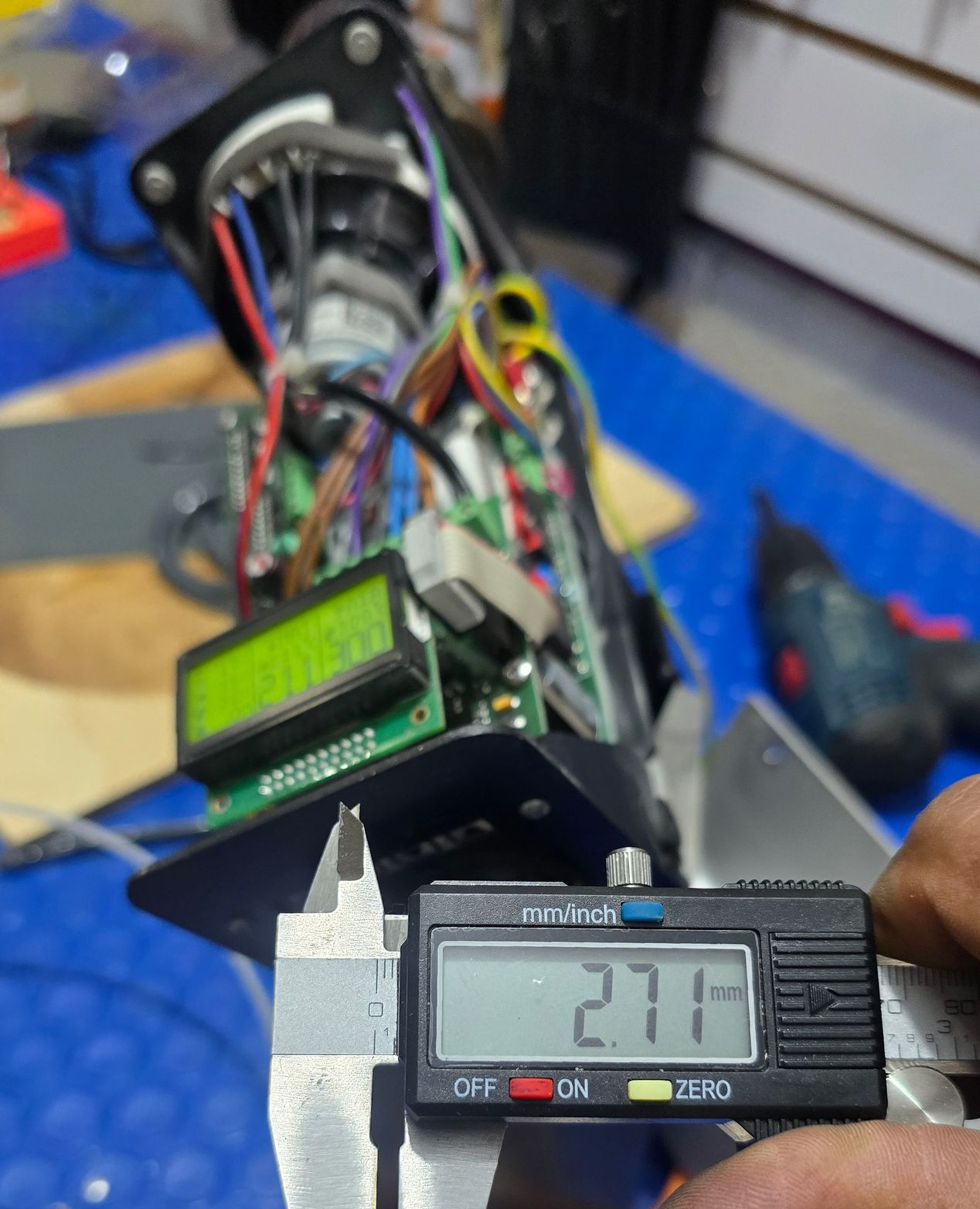 A person using digital calipers to measure a component in a device with a circuit board, display, and wires. The measurement reads 27.1 mm.