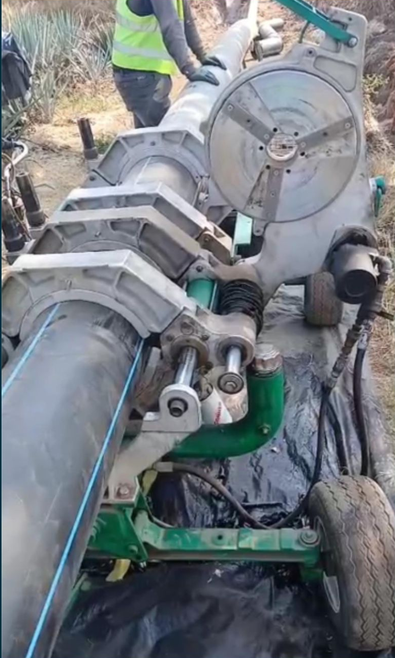 A worker using a pipe fusion machine to join two large black plastic pipes outdoors.
