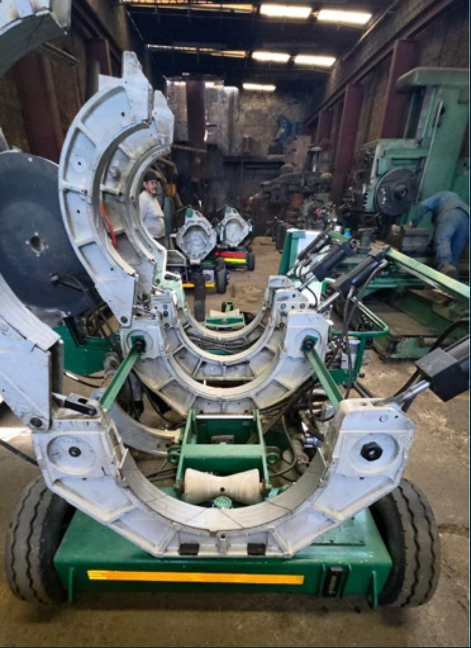 Large pipe fusion machine in a workshop. Green, white, and black machinery, workers in the background.