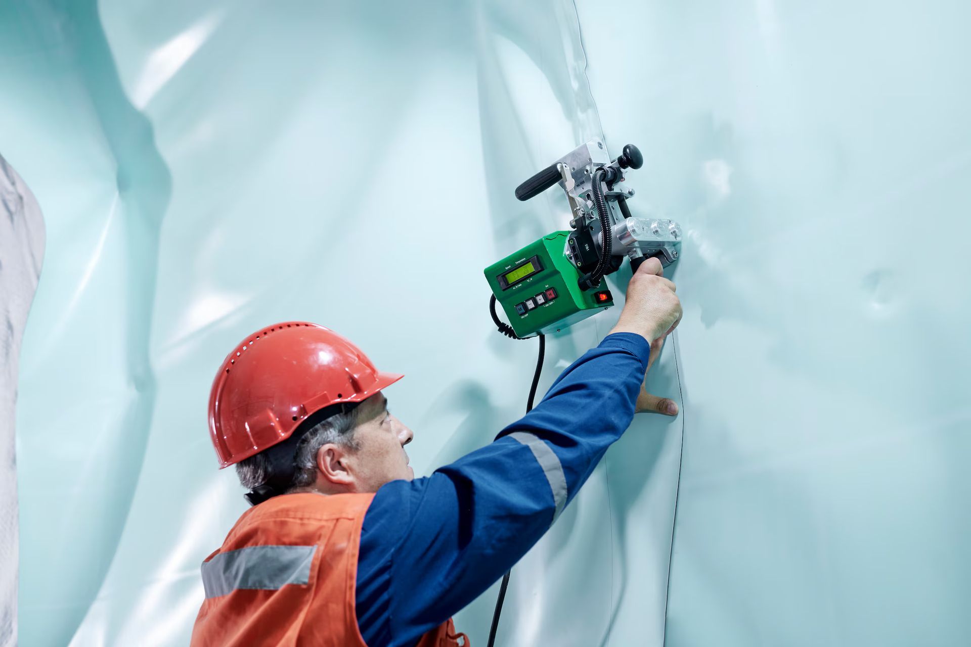 Worker in orange vest and red hard hat welding blue plastic sheet with green machine.