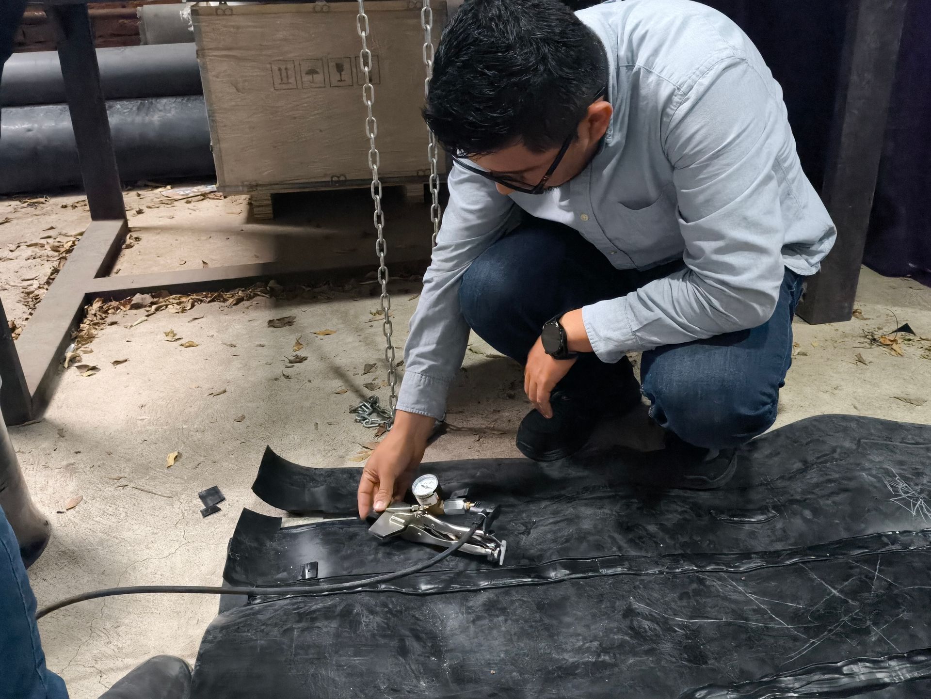 Man in blue shirt kneels, examining a device attached to black material on a floor, indoors.