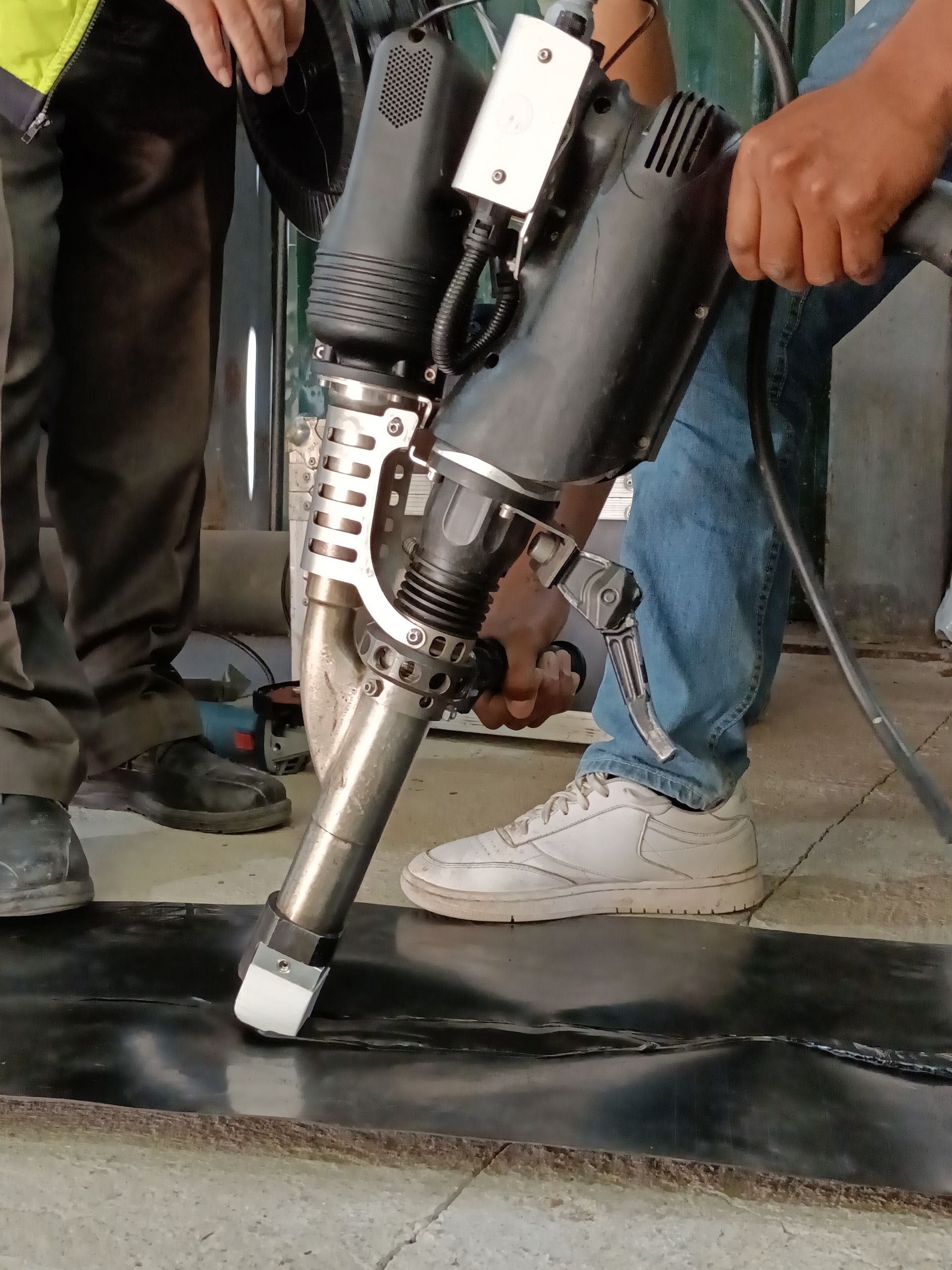 Workers use a handheld hot air welding tool to join black material on a concrete floor.