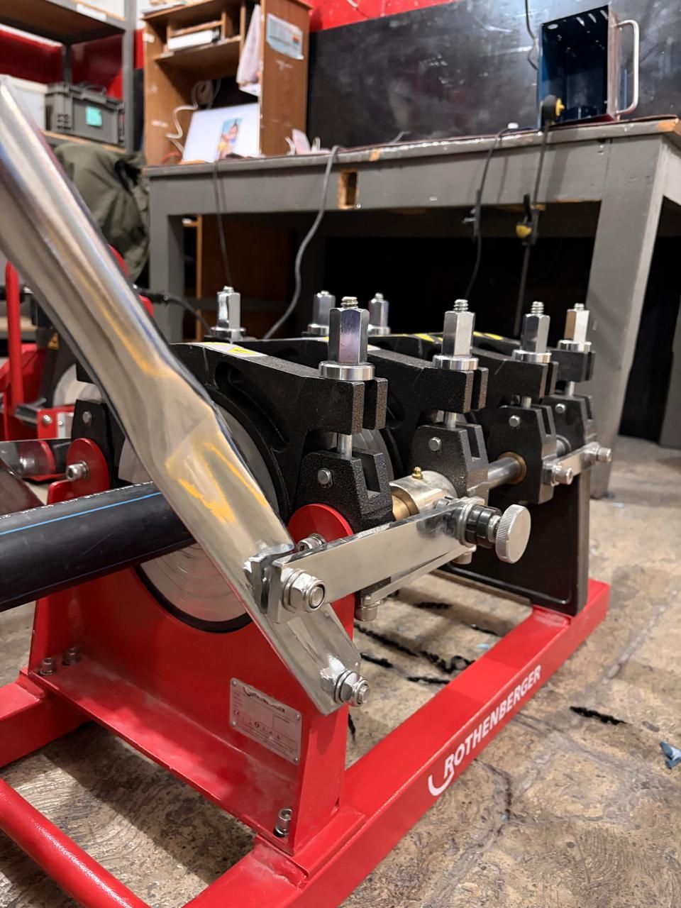 Red machine welding a metal pipe. A shop setting with tools and equipment in the background.