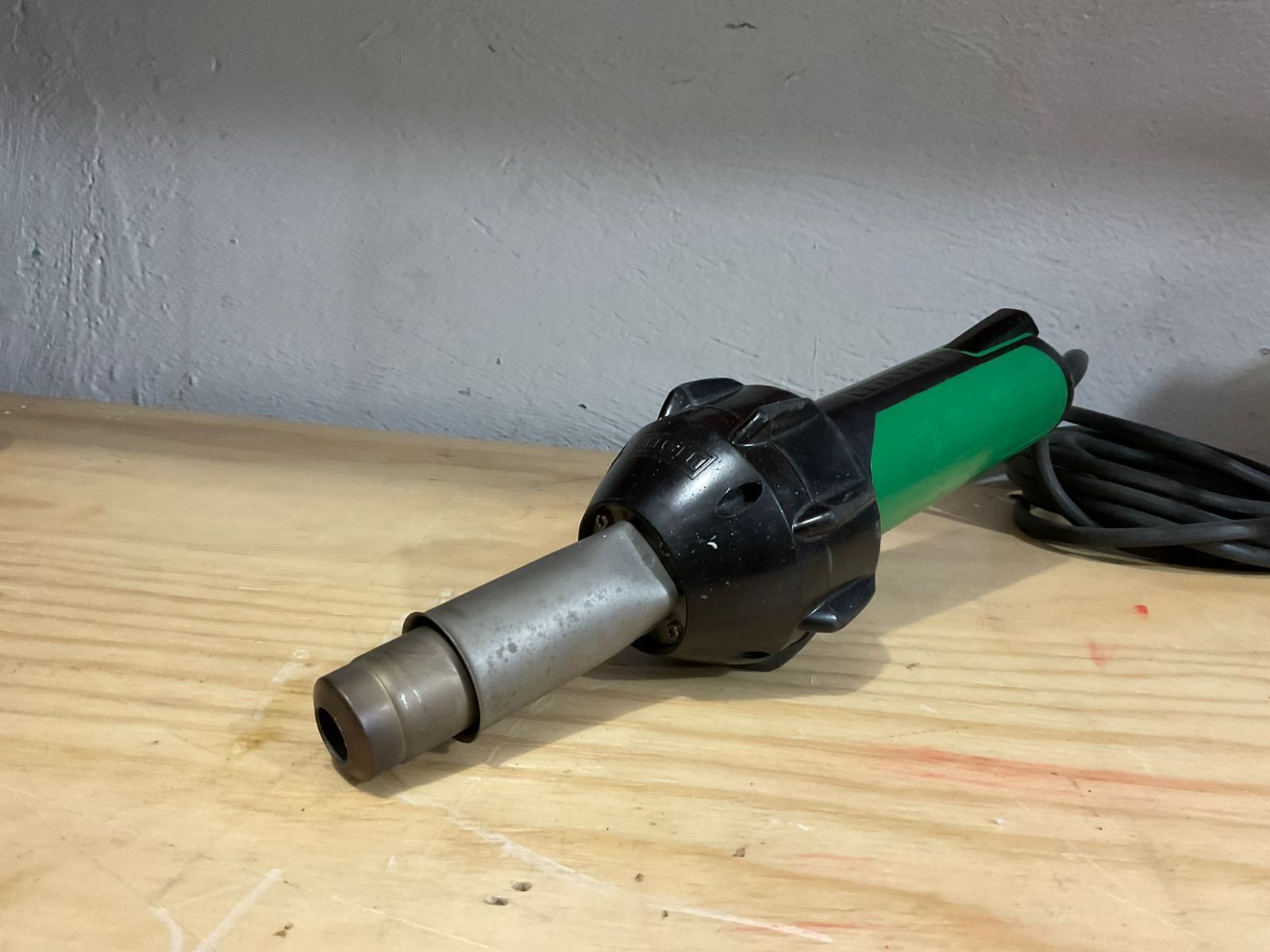 Green and black heat gun on a wooden surface, with a black cord.