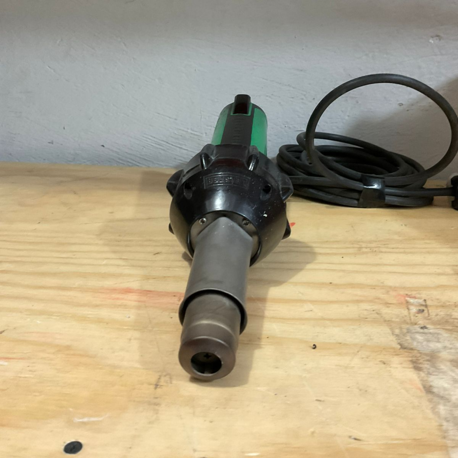 Heat gun with a green handle and black nozzle sits on a wooden surface. A black cord is coiled beside it.