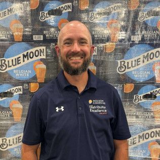 Smiling man in a navy polo stands before a Blue Moon and Belgian Moon backdrop.