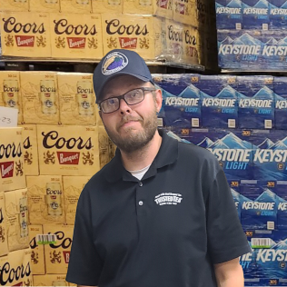 Man wearing a black shirt and blue cap standing in front of stacked Coors and Keystone beer cases